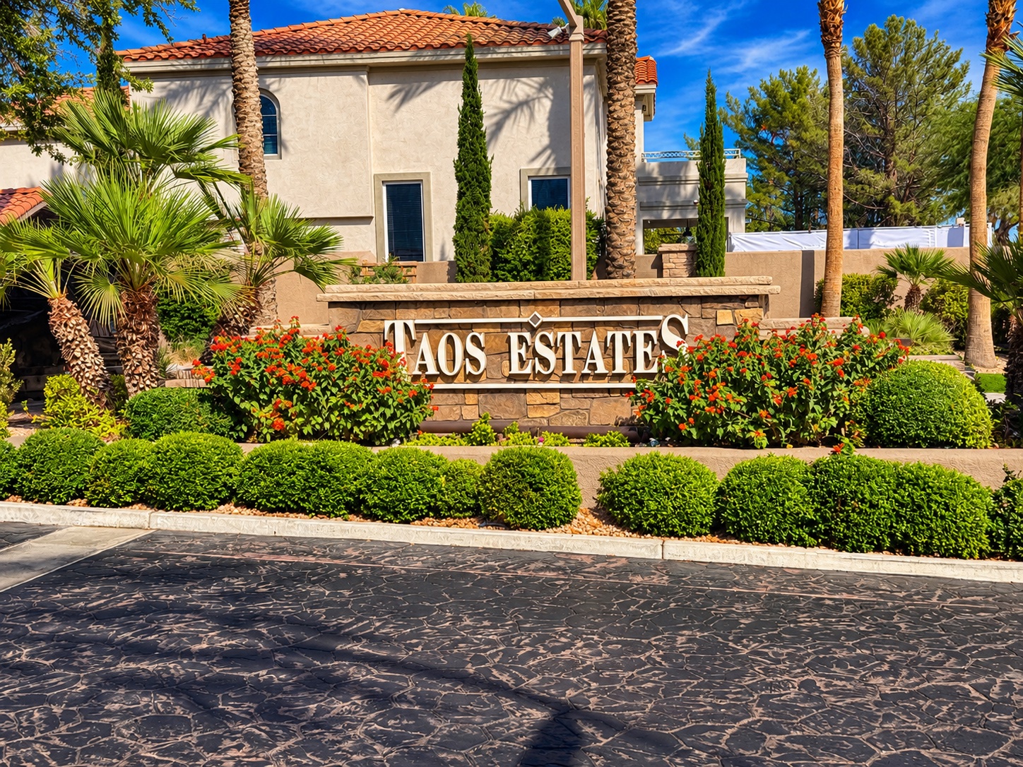 Taos Estates entrance sign with palm trees, flowering shrubs, and a tile-roof home in The Pueblo, Summerlin, Las Vegas 89128
